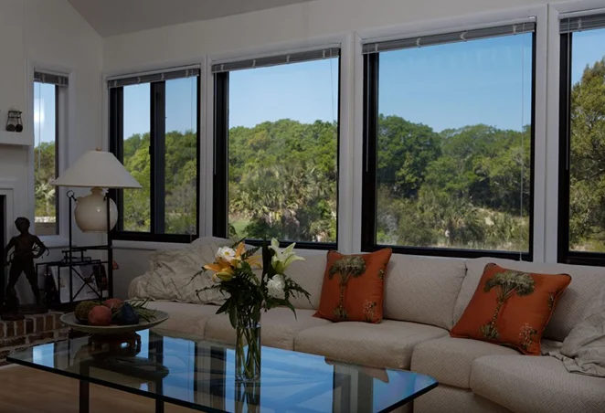 Living room with a large beige sectional sofa, glass coffee table, and wide tinted windows offering a clear view of green trees and landscape outside, reducing glare and enhancing privacy.