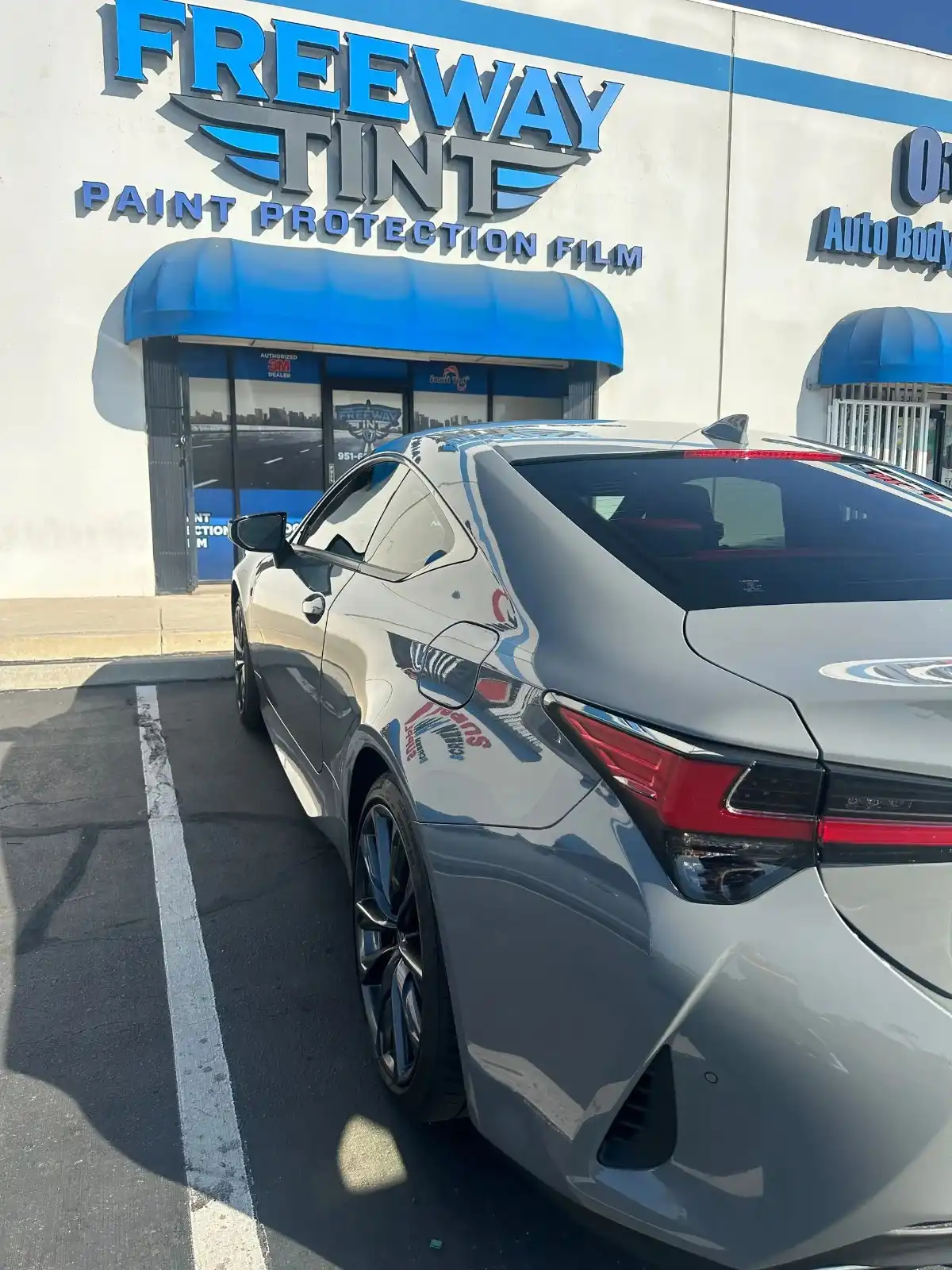 Metallic gray Lexus coupe with tinted windows parked outside Freeway Tint, with the storefront’s blue awning and “Paint Protection Film” signage visible above.