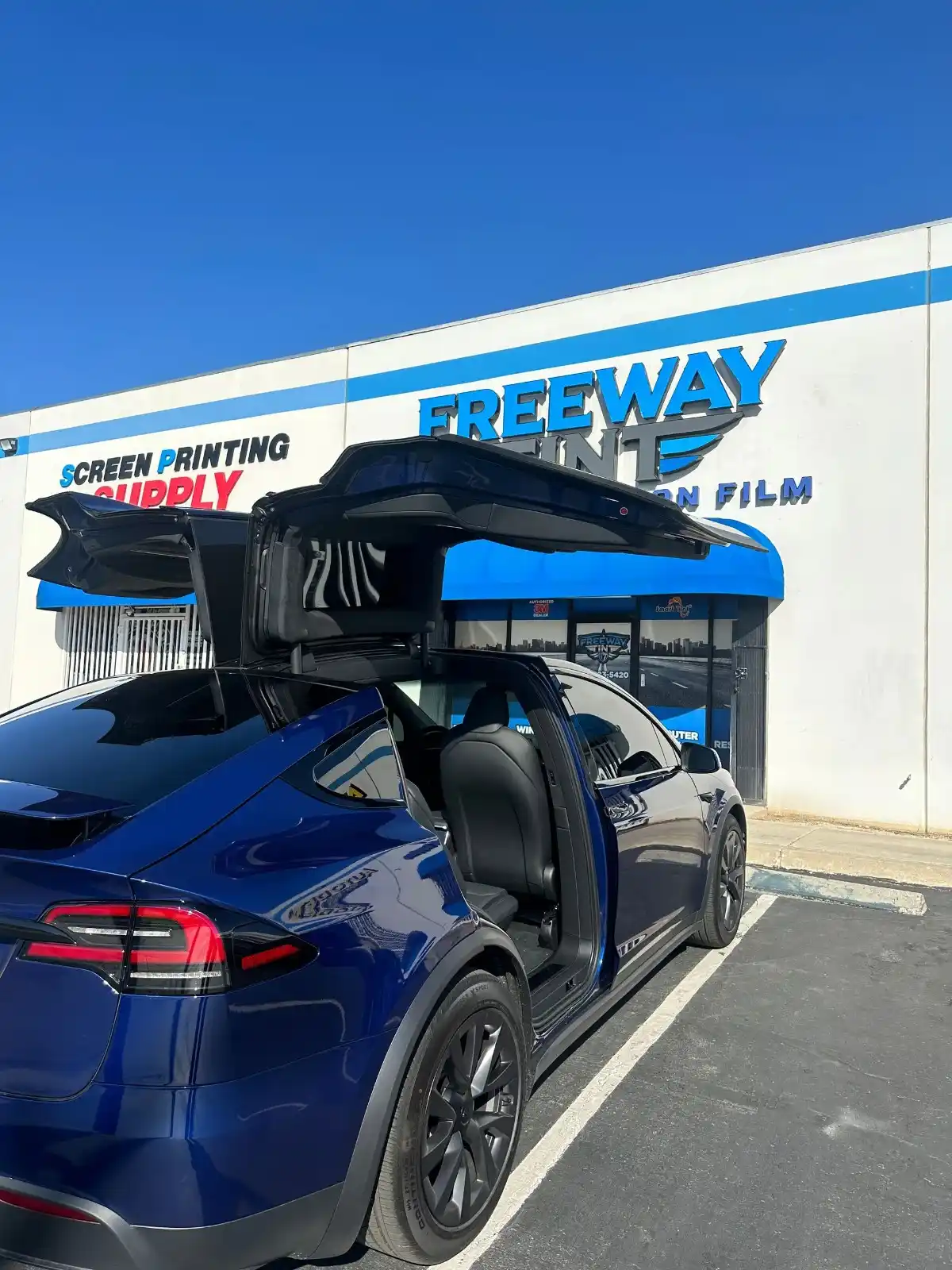 Dark blue Tesla Model X with tinted windows and both falcon-wing and front doors open, parked outside Freeway Tint under a clear blue sky.
