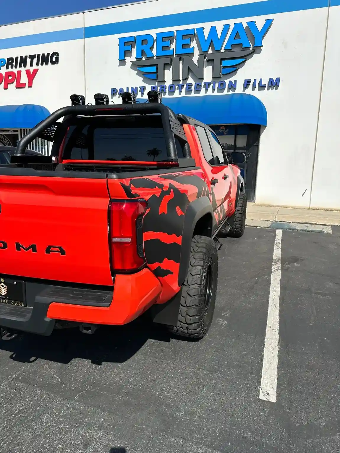Red Toyota Tacoma with black and red camo detailing, tinted windows, and off-road accessories parked outside Freeway Tint, beneath signage reading “Paint Protection Film.”