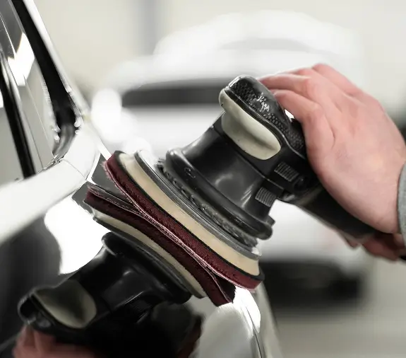 Close-up of a hand using a dual-action polisher to buff the surface of a black vehicle, creating a glossy, reflective finish.