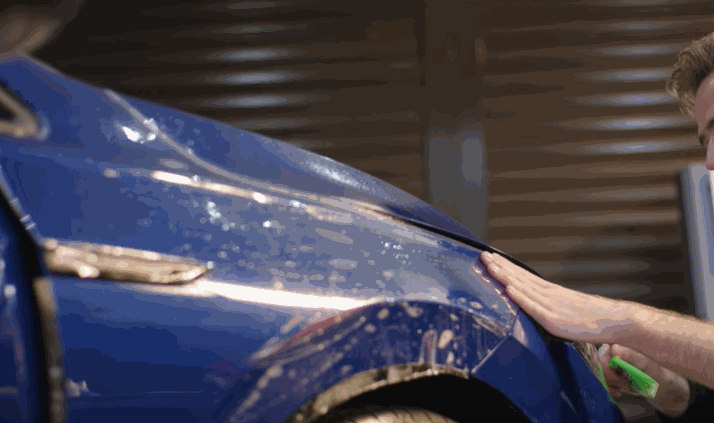A person presses paint protection film onto a blue car fender, using their hand to flatten the material over the curved surface. Water droplets and reflections highlight the glossy finish as the film settles into place.