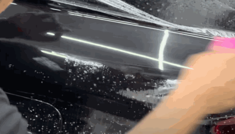 A close up of a black car panel covered in soapy water as a person smooths paint protection film across the surface. Small bubbles and streaks are visible under the film as it is carefully pressed down to adhere evenly.
