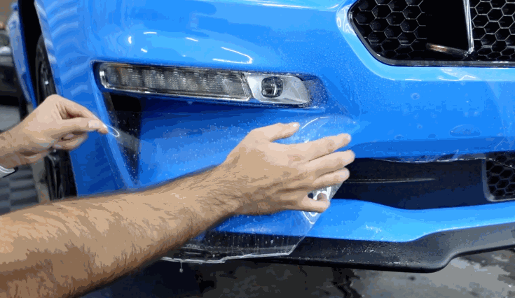 Installer positions clear paint protection film over the front bumper of a blue car and works it around the grille and light opening.
